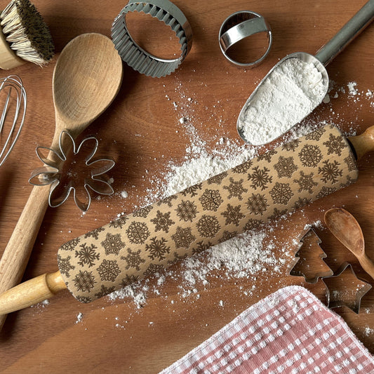 Baking tools including a rolling pin with embossed designs, wooden spoons, and cookie cutters on a wooden surface with flour.