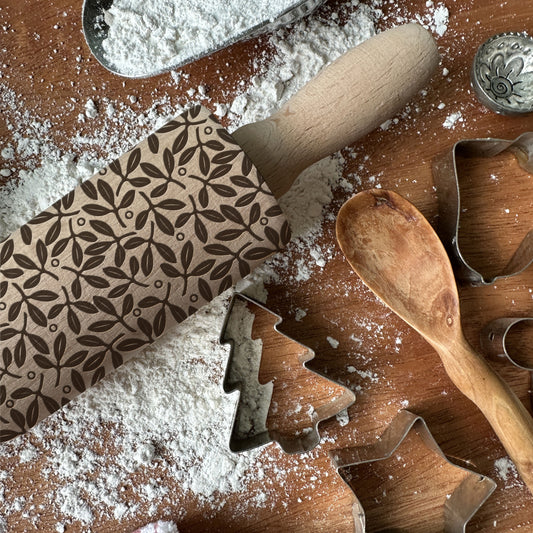 Wooden rolling pin with embossed pattern on a flour-dusted surface with cookie cutters and a wooden spoon.