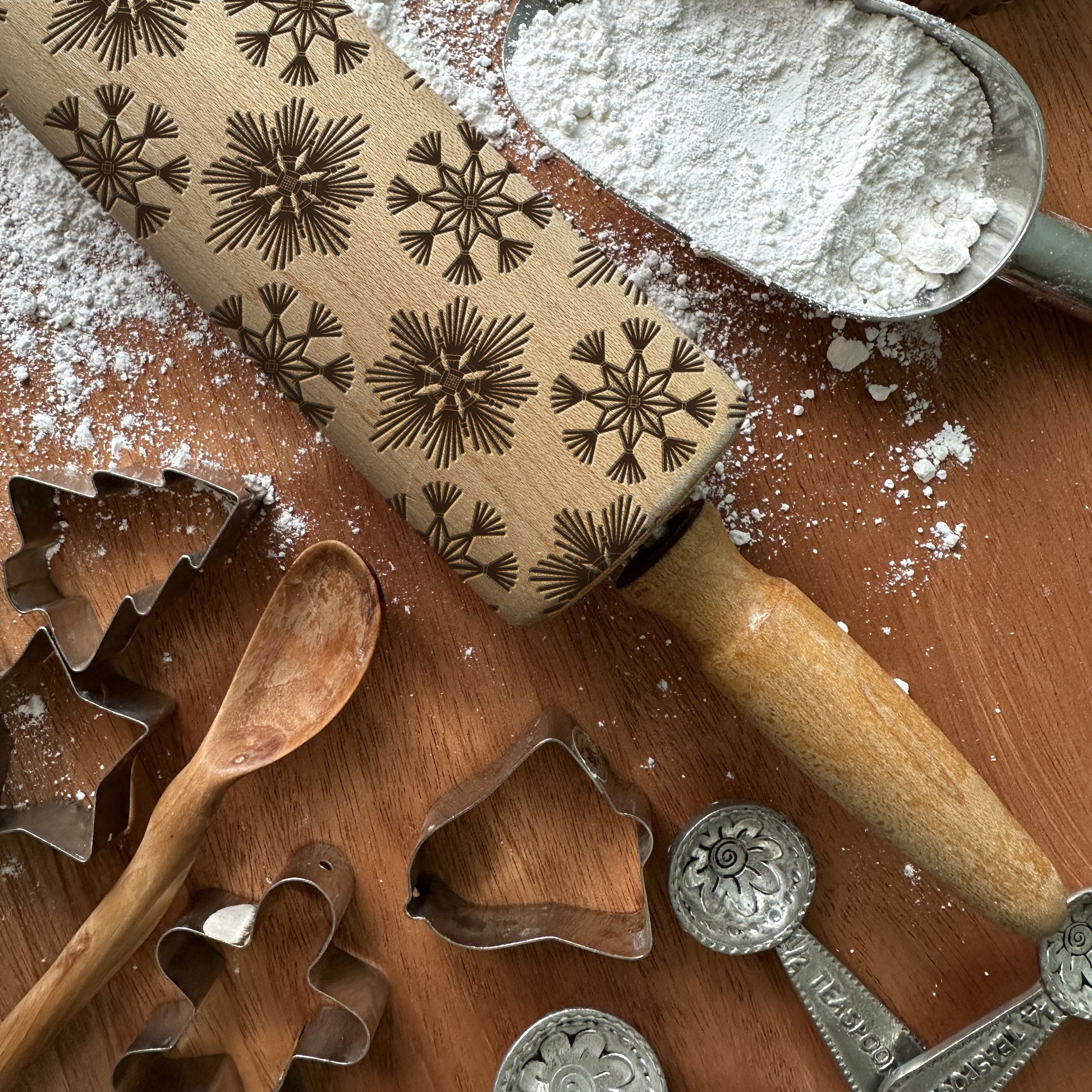 Rolling pin with snowflake patterns on a wooden surface with cookie cutters and flour.