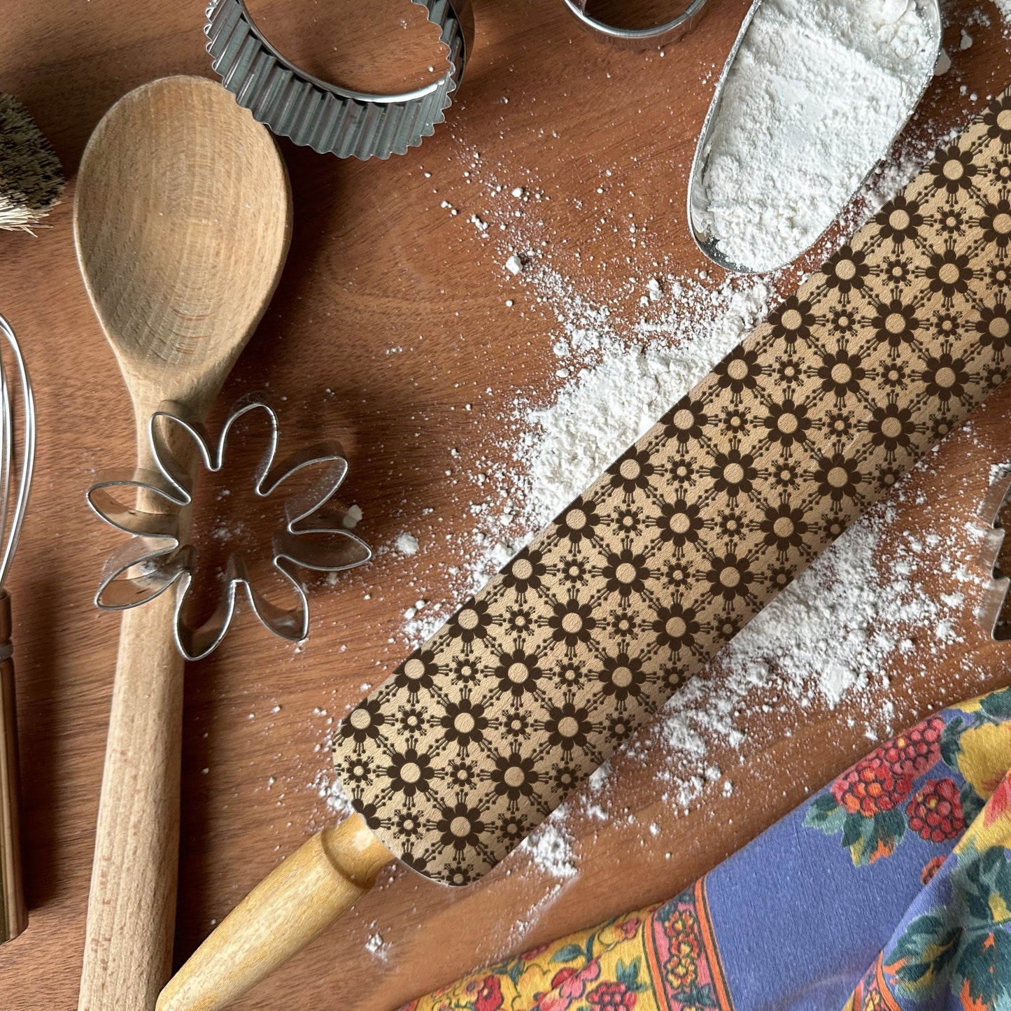 Baking tools including a wooden spoon, rolling pin with floral design, and cookie cutters on a wooden surface with flour.