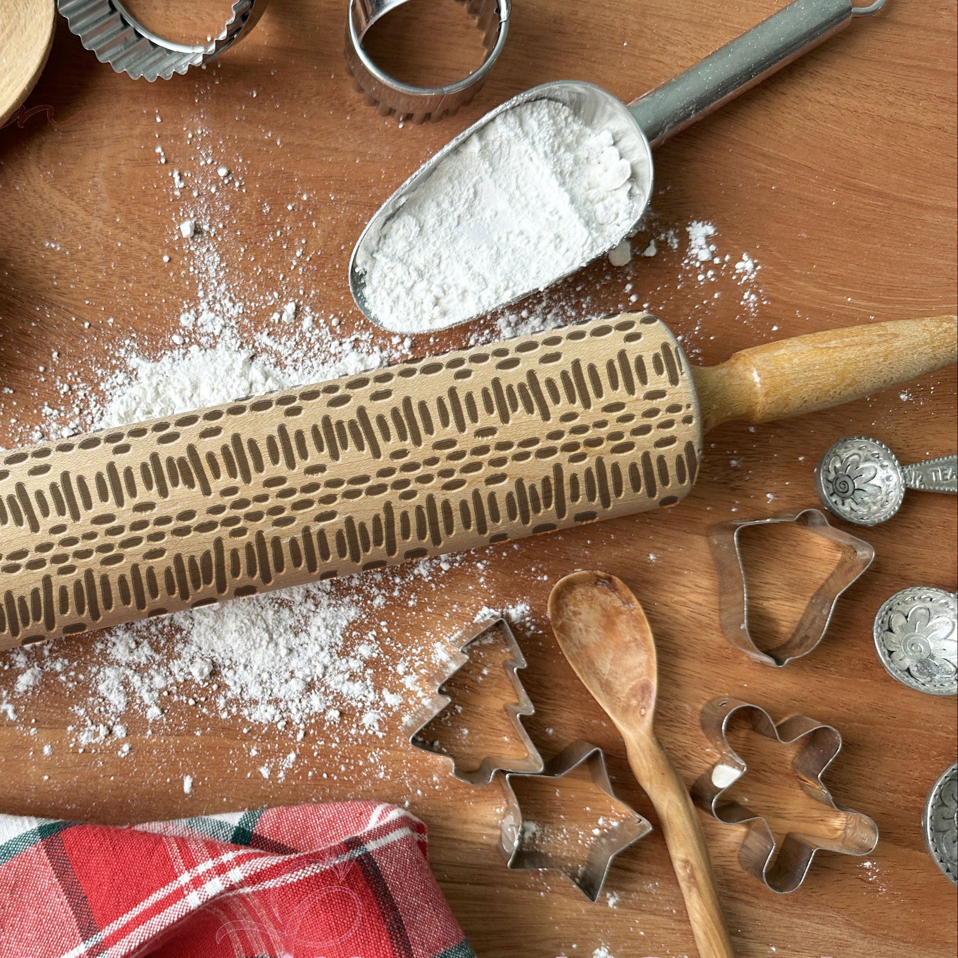 Wooden rolling pin with embossed patterns on a wooden surface with flour, cookie cutters, and a wooden spoon.