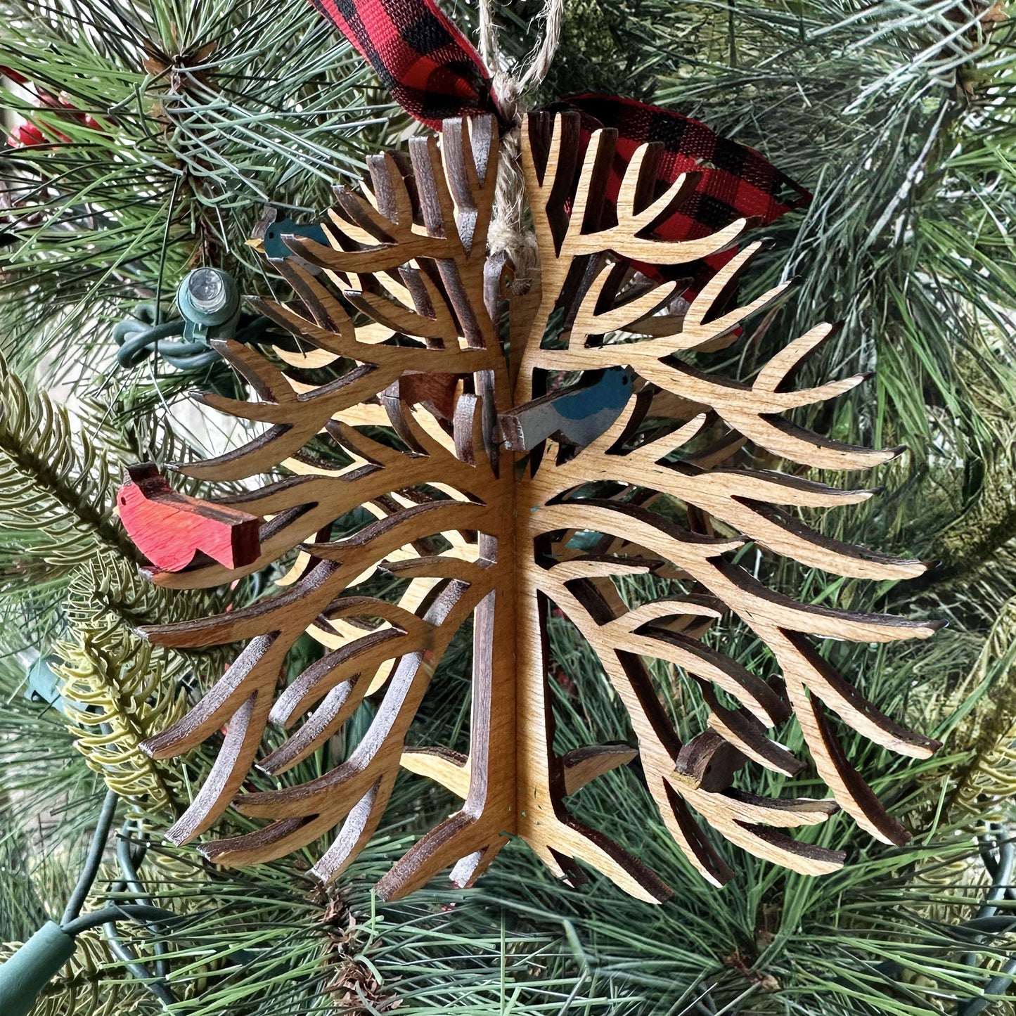 Decorative wooden ornament with a bird on a tree branch, hanging from a red and black plaid ribbon, against a Christmas tree background.