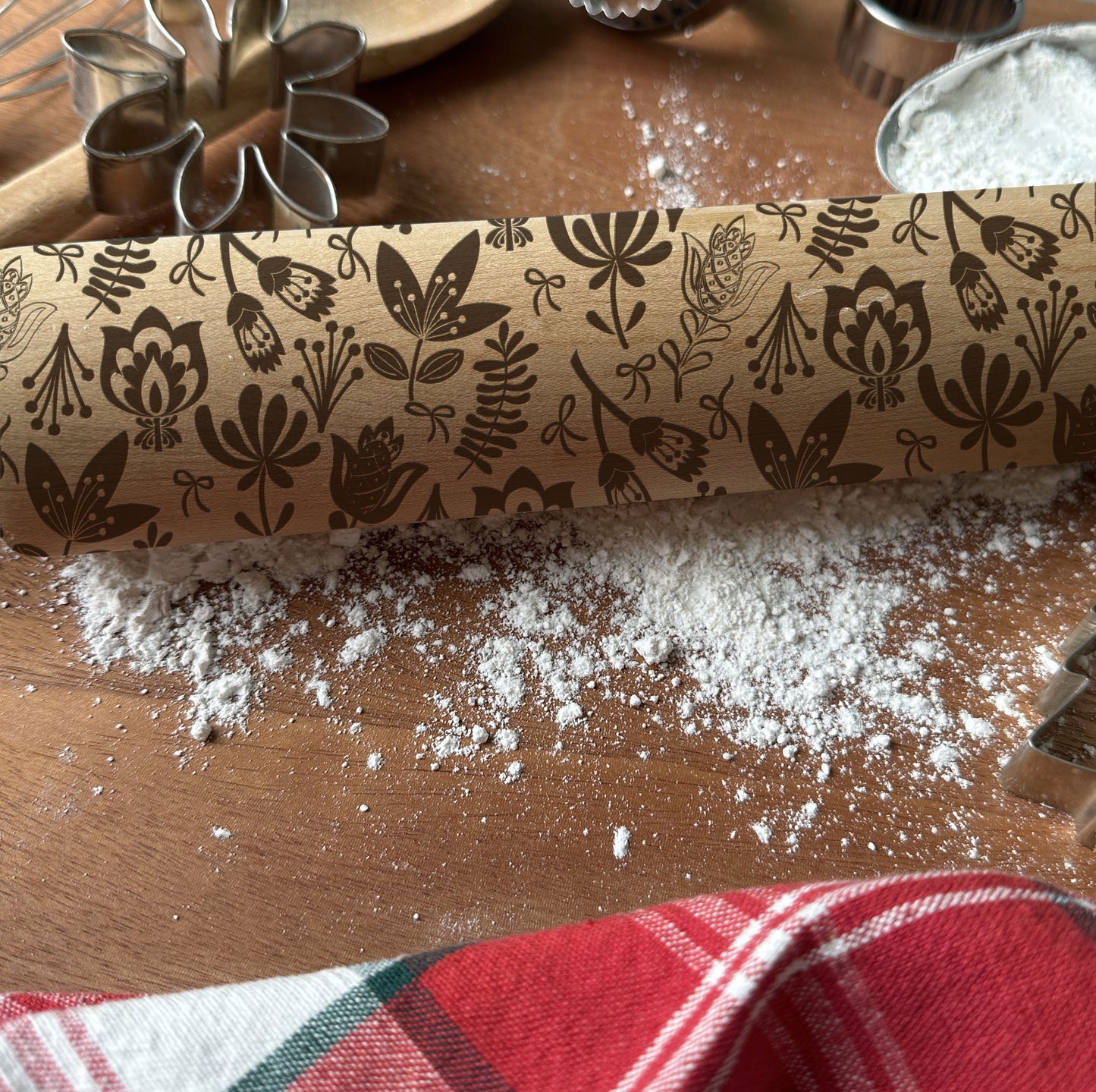 Decorative rolling pin with floral patterns on a wooden surface with flour and cookie cutters.