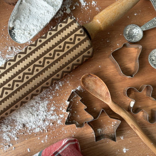 Wooden rolling pin with pattern, metal cookie cutters, and flour on a wooden surface.