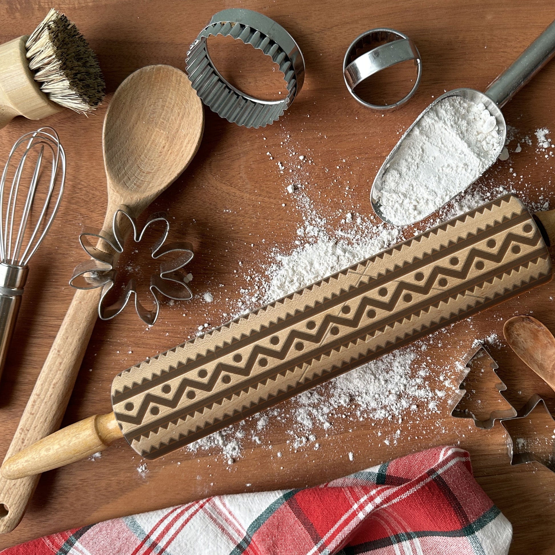 Baking tools including a rolling pin with a decorative pattern, wooden spoon, whisk, and cookie cutters on a wooden surface.